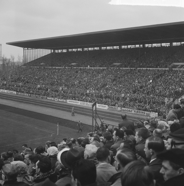 Schwarzes und weißes Foto eines vollen Stadions mit Zuschauern, die ein Fußballspiel beobachten, einige stehen und andere sitzen auf den Tribünen, mit Bannern, Pfählen, einer Hütte, Bäumen, einem Turm und einem bewölkten Himmel im Hintergrund.