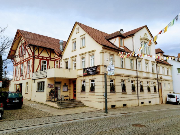 Großes modernes Gebäude mit der Aufschrift "Stadtbahnhof" in Baden-Württemberg, mit vielen Fenstern, umgeben von Bäumen, Straßenmöbeln, Fahrzeugen, Fußgängern und Werbetafeln unter einem bewölkten Himmel.