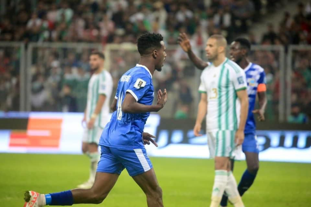 Ein Fußballspieler in einem blauen und weißen Trikot feiert mit erhobenen Armen, umgeben von einer Gruppe in weißen und grünen Trikots, mit einem unscharfen Hintergrund, der ein weißes und blaues Board und ein Geländer zeigt.