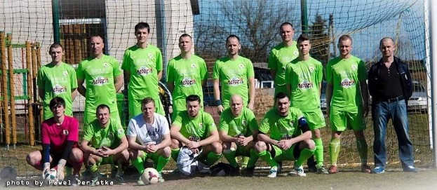Eine Gruppe von Männern in grünen T-Shirts steht auf einem Fußballfeld, einige in der Hocke, mit einem Ball vor ihnen in der Nähe eines Torpfostens, Bäume, Gebäude und Fahrzeuge im Hintergrund.