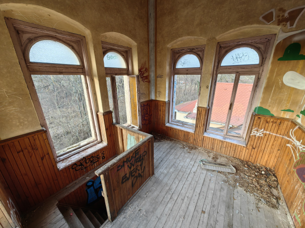 Verlassenes Gebäude mit Graffiti an den Wänden und Fenstern, Holzboden, Treppe, verstreuten Gegenständen und Bäumen mit einem Haus im Hintergrund.