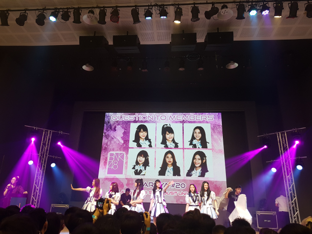 Eine Gruppe von Menschen performs auf einer hell erleuchteten Bühne mit Trägern, während eine Menge darunter Fotos macht; ein großes Display im Hintergrund zeigt Bilder der K-Pop-Gruppe BK48.