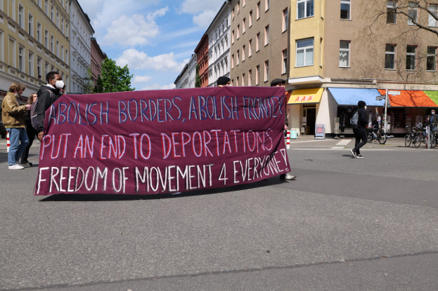 Eine Gruppe von Menschen marschiert auf einer Straße und hält ein Banner mit der Aufschrift "Abolish Borders, Abolish Frontiers, Put an End to Deportations, Freedom of Movement 4 Everyone", mit Gebäuden, Bäumen und Fahrrädern, die die Straße säumen, unter einem bewölkten Himmel.