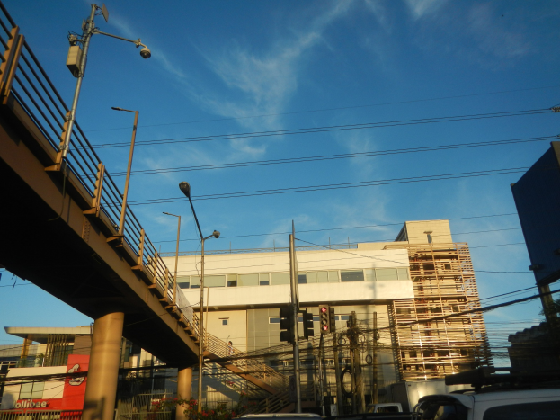 Stadtstraße mit parkenden Autos, eine Brücke mit Geländern, Laternen, Strommasten mit Drähten, Verkehrsampeln, Gebäude mit Fenstern und ein bewölkter Himmel im Hintergrund.