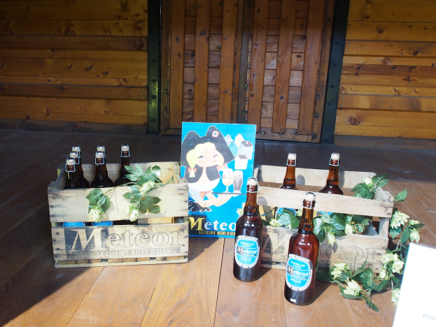 Ein Holzkasten voller Bierflaschen auf einem Holzboden in einer Gartenumgebung, mit Blättern und Blumen drumherum und einer Holzwand und einer Tür im Hintergrund.