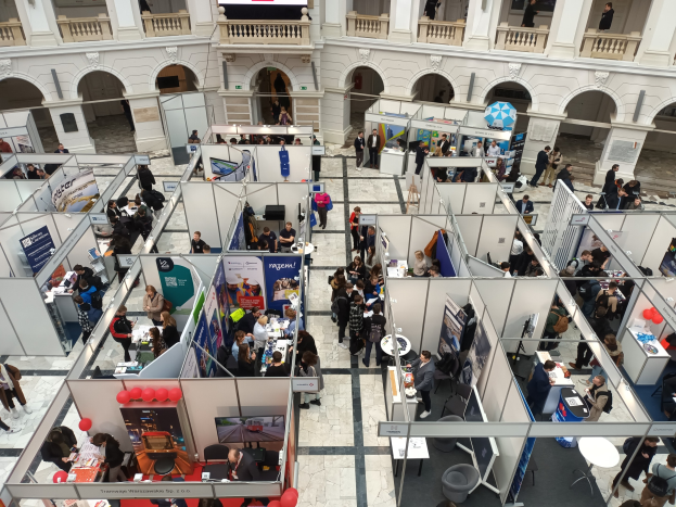 Große Menschenmenge auf einer internationalen Fachmesse mit Ständen, Bannern, Tischen, Stühlen, Ballons und einem Gebäude mit Bögen, Säulen, Geländern und einem Bildschirm im Hintergrund.