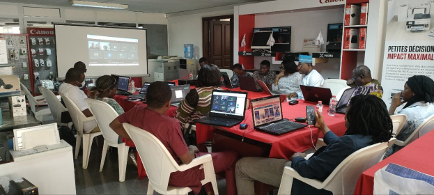 Gruppe von Menschen an einem Tisch mit Laptops, Flaschen und anderen Gegenständen während eines digitalen Marketing-Workshops, mit einem Banner, einem Bildschirm, einer Tür und Deckenleuchten im Hintergrund.