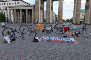Eine Gruppe von Fahrr├Ądern, die vor dem Brandenburger Tor in Berlin, Deutschland, geparkt sind, mit verstreuten Taschen und Gegenst├Ąnden, einer Fahne, Geb├Ąuden, B├Ąumen, Masten, Flaggen und einem klaren blauen Himmel im Hintergrund.