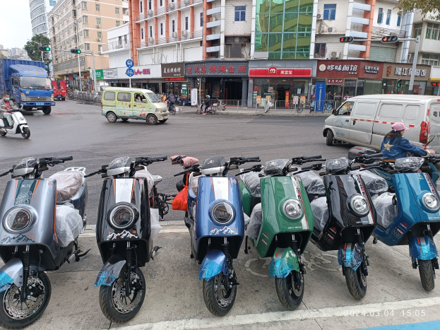 Eine Reihe von E-Scootern, die auf einer Straße geparkt sind, mit einer Person in einer Mütze davor, vor einem Hintergrund aus Gebäuden, Verkehrsampeln, Bäumen und einem klaren Himmel.