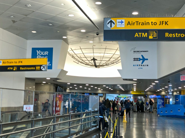 Eine Gruppe von Menschen geht durch eine Flughafenhalle mit einer Rolltreppe auf der linken Seite, Schilder an den Wänden und einer Tafel mit der Aufschrift "Airtrain nach JFK".