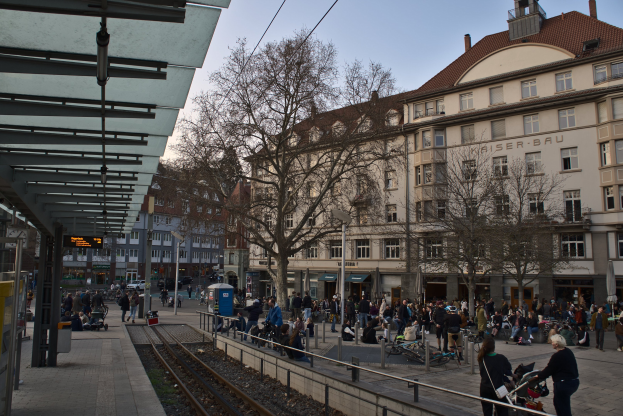 Eine belebte Straßenkreuzung mit einem Bahnhof auf der linken Seite, Menschen gehen, sitzen auf Bänken und stehen in der Nähe, umgeben von Gebäuden, Bäumen, Laternenmasten und Texttafeln, unter einem sichtbaren Himmel.