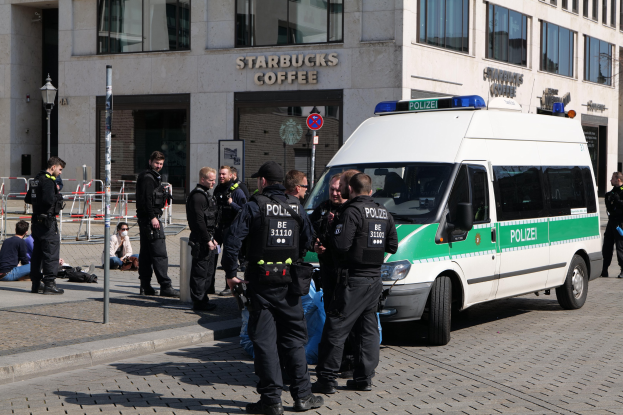 Eine Gruppe von Polizisten steht vor einem Starbucks Kaffeehaus, rechts ein Van und links einige Personen; im Hintergrund sind ein Gebäude, ein Schild, ein Laternenpfahl und ein Zaun zu sehen.