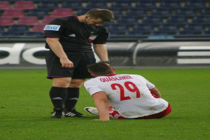 Ein Fuballspieler sitzt neben einem Schiedsrichter auf dem Boden in einem Stadion, beide tragen Sportkleidung, mit Brettern und Stühlen im Hintergrund.