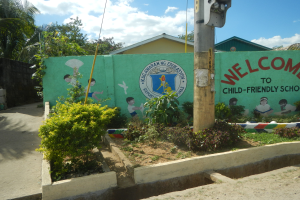 Eine grüne Wand mit einem Schild, auf dem "Willkommen in der kinderfreundlichen Schule" steht, umgeben von Häusern, Bäumen, Pflanzen und einem Mast mit einer Lampe, unter einem bewölkten Himmel.