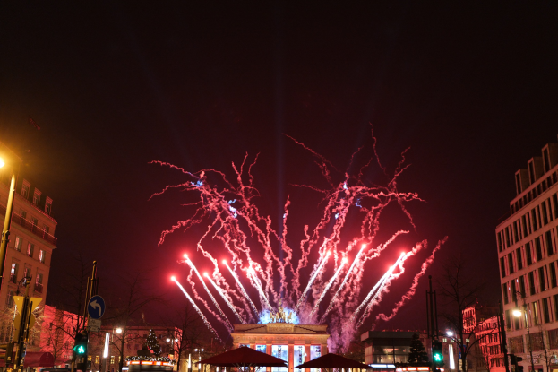 Eine belebte Straße in Berlin an Silvester, voller Menschen, Fahrzeuge und Gebäude, erleuchtet von Feuerwerk und Gebäudebelichtung, die eine festliche Atmosphäre schaffen.