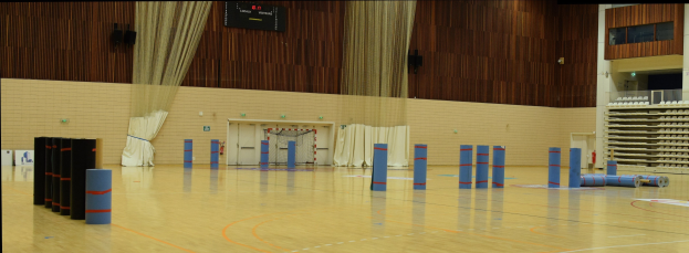 Großer Turnsaal mit Basketballfeld, Regalen auf der rechten Seite, Vorhängen und Leinwand an der Rückwand und sichtbarem Boden.