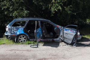 Ein beschädigt aussehendes Auto am Rödrand mit Gras und Bäumen im Hintergrund.
