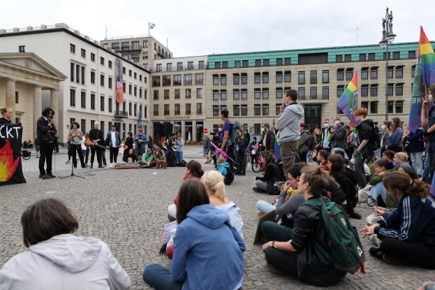 Eine Gruppe von Menschen, die auf dem Boden vor einer Menge sitzen, die Fahnen und Transparente hält, während einer Anti-Schwulen-Demo in Berlin. Im Hintergrund sind eine Statue, Gebäude und ein Mikrofonständer zu sehen.