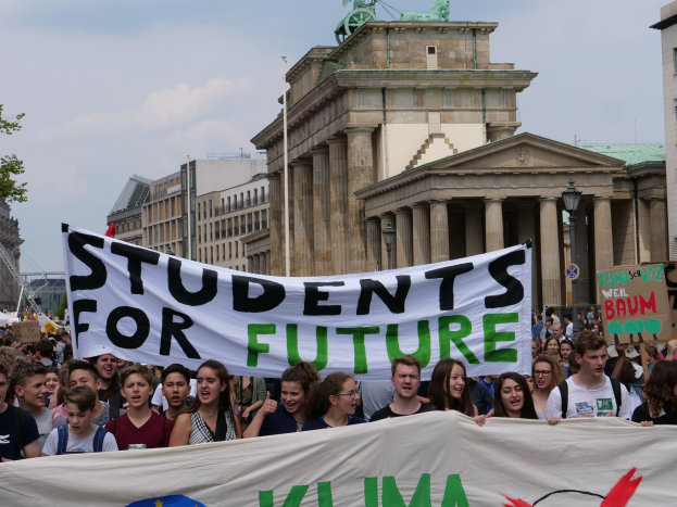 Gruppe von Schülerinnen und Schülern marschiert in Berlin mit einem bunten "Students for Future"-Schild an Gebäuden, Bäumen und Himmel vorbei.