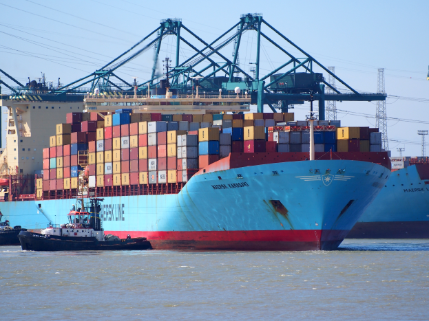 Ein großes Containerschiff liegt neben einem Schlepper, mit vielen Containern an Bord und Türmen und Drähten im Hintergrund bei klarem blauem Himmel.