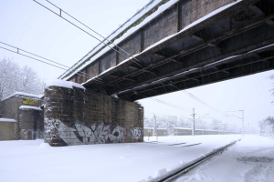 Eine schneebedeckte Bahnschiene unter einer Brücke mit Graffiti, Strommasten mit Drähten, Bäumen und einem Himmel im Hintergrund.