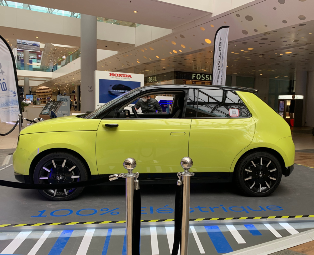 Gelbes wasserstoffbetriebenes Elektroauto vor einem Gebäude mit Absperrpfosten, Bannern, Menschen, Schildern, einer Topfpflanze, Deckenleuchten und Lüftern.