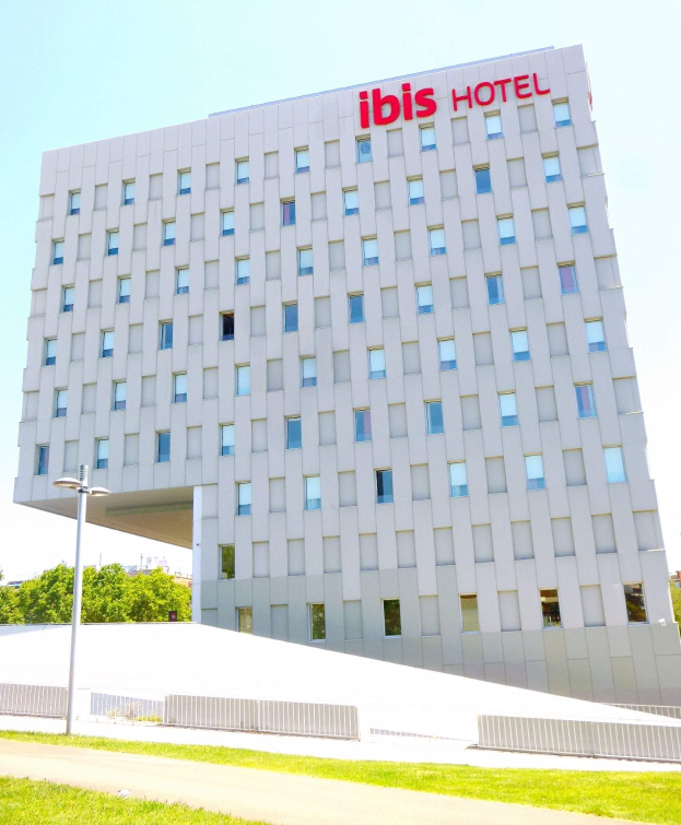 Großes Gebäude des Ibis Hotels in Berlin mit Fenstern, einer Straßenlaterne, Bäumen, einem Zaun und einem Weg unter einem bewölkten Himmel.