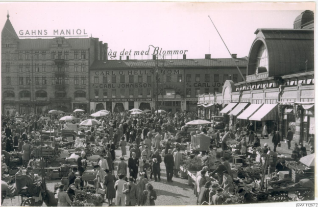 Schwarzes und weißes Foto eines belebten Berliner Markts mit Menschen, Gemüsekarren, Schirmen und Gebäuden im Hintergrund.