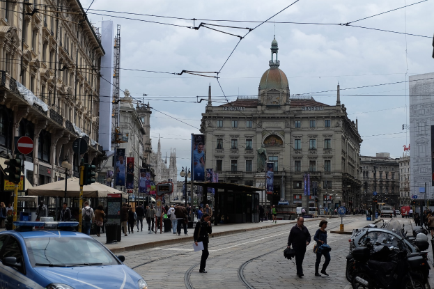 Eine Straße mit einem parkenden Polizeiwagen, Führer mit Taschen, fahrende Fahrzeuge, Gebäude mit Fenstern und Plakaten, Laternen, Verkehrssignale und ein bewölktes Himmel.