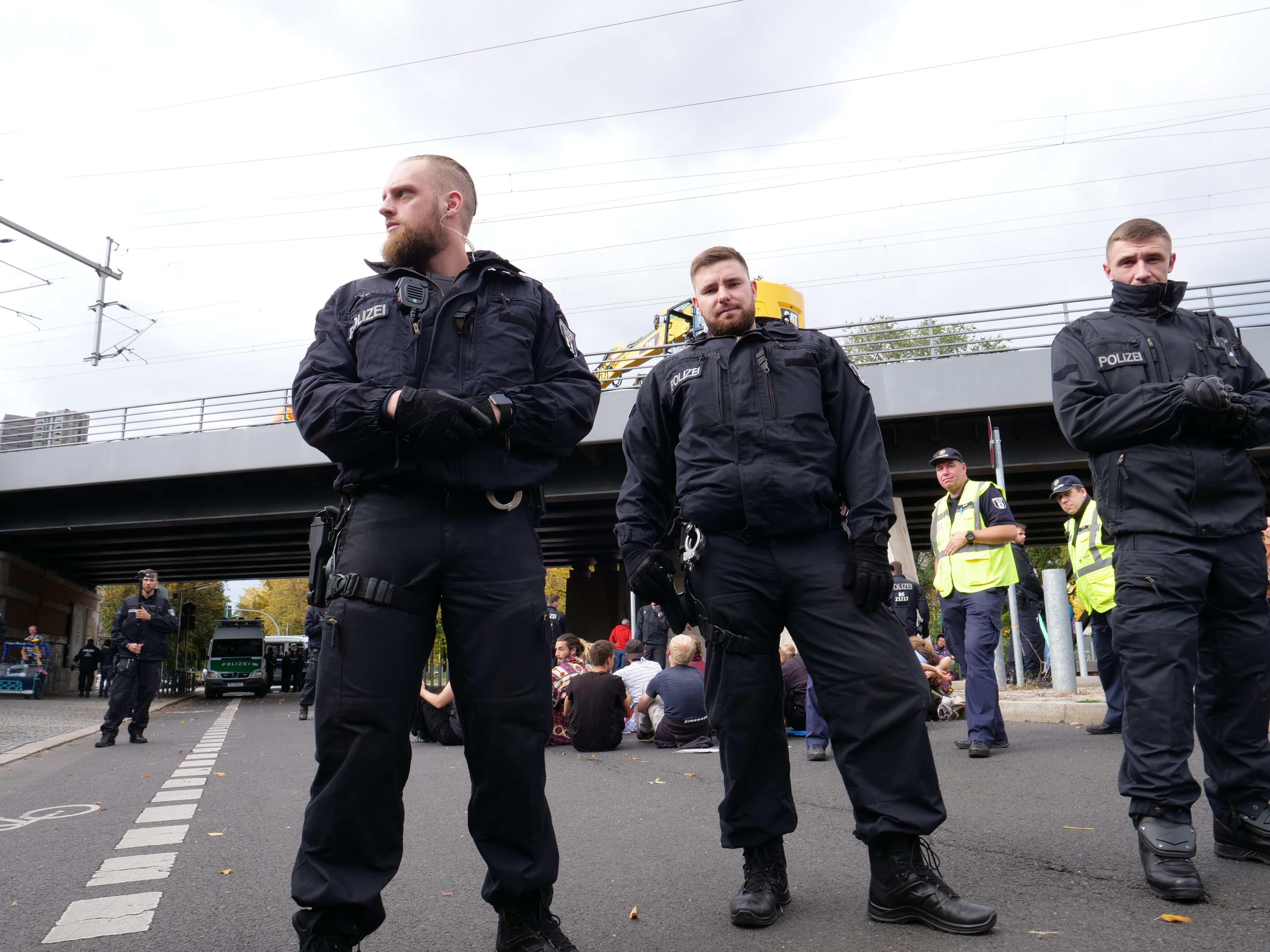 Eine Gruppe von Polizisten steht an der Straßenseite mit Menschen, Fahrzeugen, einer Brücke, Bäumen, Masten, Drähten und einem klaren blauen Himmel im Hintergrund.