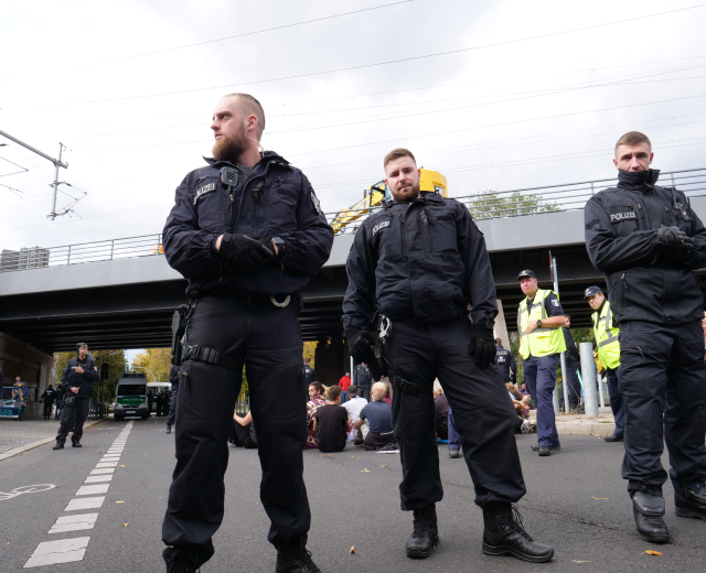 Eine Gruppe von Polizisten steht an der Straßenseite mit Menschen, Fahrzeugen, einer Brücke, Bäumen, Masten, Drähten und einem klaren blauen Himmel im Hintergrund.