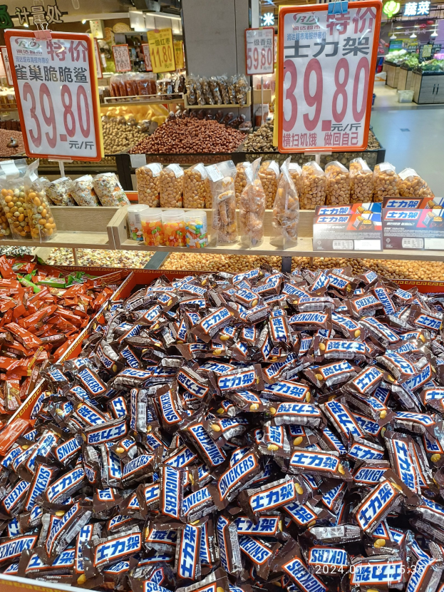 Vitrine voller verschiedener Süßigkeiten, darunter Snickers-Riegel, in einem Geschäft mit Lebensmitteln auf Regalen und Deckenlampen im Hintergrund.