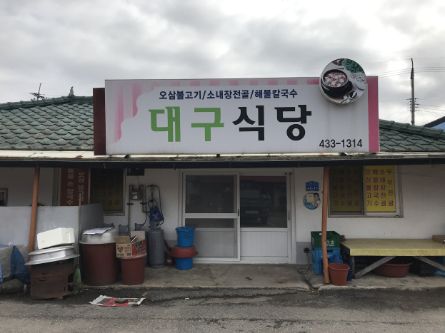 Kleines Gebäude mit einer Tafel, die "Koreanisches Restaurant" lautet, einer Tür, Fenstern, Behältern, Eimern, einem Tisch, Pfählen, Drähten, einem weiteren Haus und einem bewölkten Himmel.