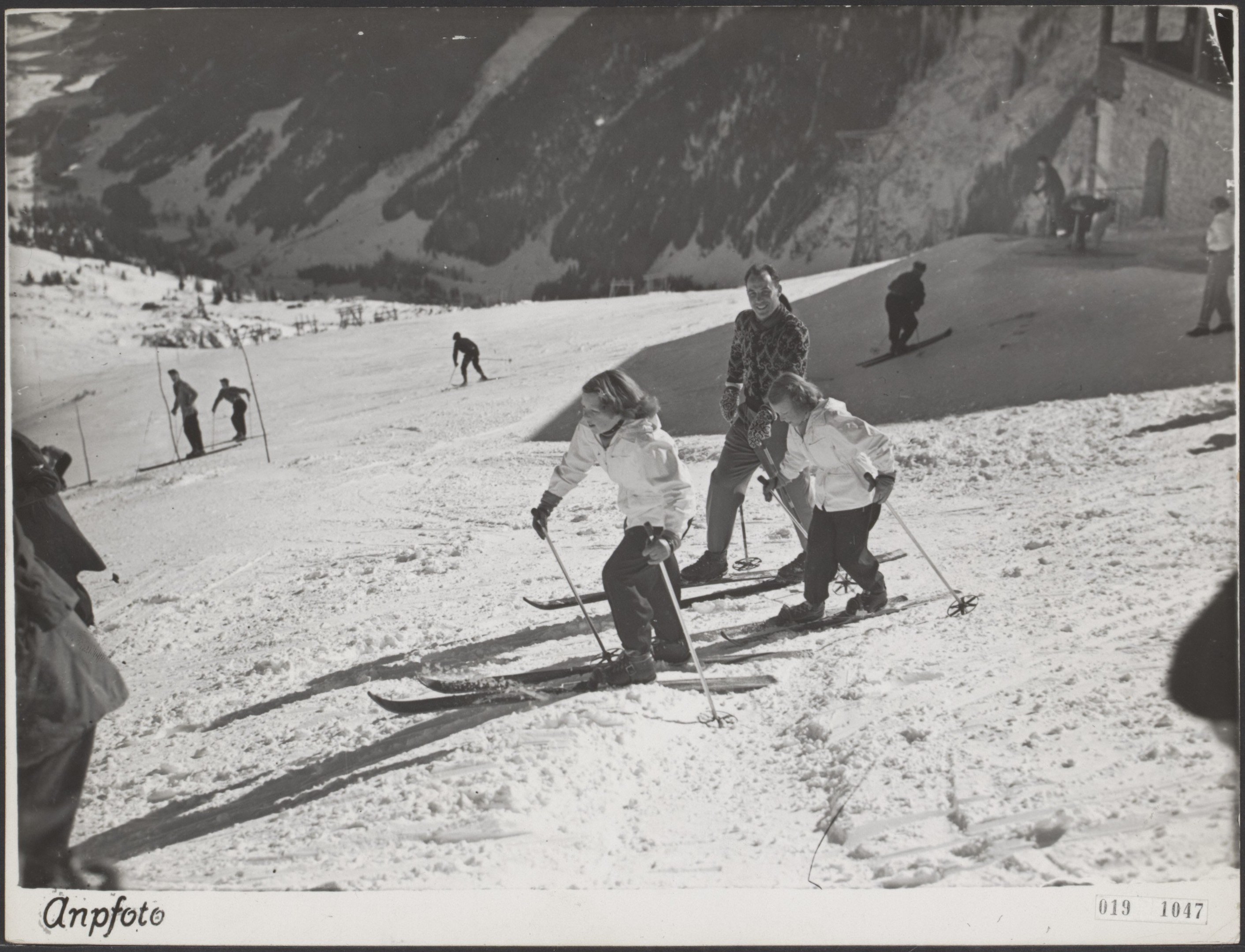 Gruppe von Menschen, die eine schneebedeckte Abfahrt hinunterskifahren, jeder hält Ski-Stöcke in der Hand, mit Hügeln und einem Gebäude im Hintergrund und Text unten.