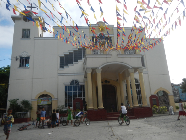 Eine Gruppe von Menschen, die vor einer weißen Kirche mit architektonischen Details wie Fenstern, Säulen, Bögen, Türen und einem Kreuzsymbol Fahrrad fahren, mit Pflanzen, einem Zaun, einer Fahne, Bäumen und einem bewölkten Himmel im Hintergrund und bunten Fahnen, die an einem Seil oben befestigt sind.