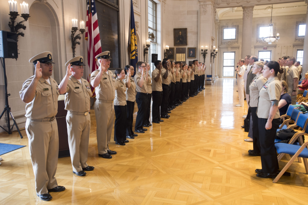 Gruppe uniformierter Menschen, die bei einer Militär-Offizier-Einführungszeremonie salutieren, in einem Raum mit Stühlen, Fotorahmen, Kerzen, einem Lautsprecher und einer Fahne.