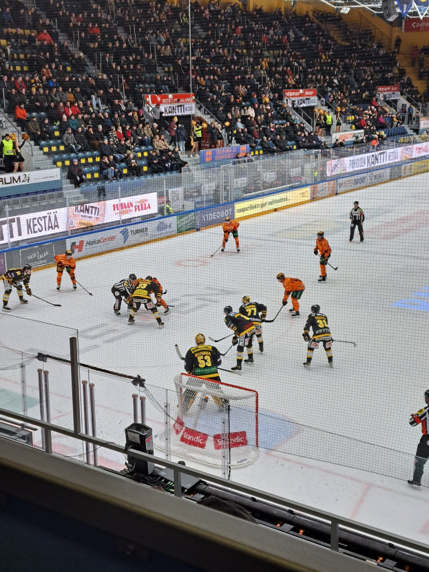 Eine Gruppe von Menschen, die Hockey auf einem Eisring mit einem Netz und einer Umzäunung spielt, mit Zuschauern im Hintergrund und Bannern mit Text, unter hellen Stadionlichtern.
