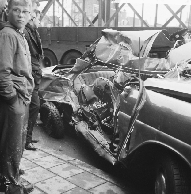 Schwarz-weißes Bild eines verbeulten Autos am Straßenrand mit Menschen in der Nähe, einem Lkw und einer Brücke im Hintergrund.