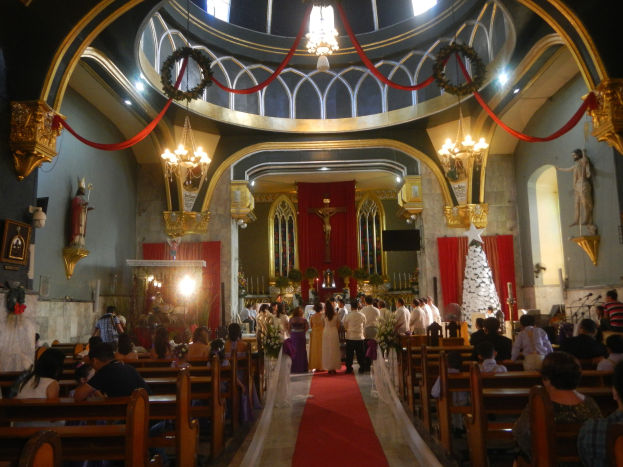 Eine Hochzeitszeremonie in einer Kirche mit einem roten Teppichgang, Sitzplätzen auf Bänken und einigen Stehenden mit Blumenbouquets, geschmückt mit Statuen, Lichtern, Kronleuchtern und anderen Dekorationen.
