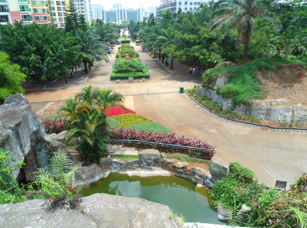Luftaufnahme eines Parks mit einem Teich, umgeben von Felsen, Pflanzen, Bäumen und Blumen, Menschen, die herumlaufen, Gebäude im Hintergrund und einem klaren blauen Himmel.