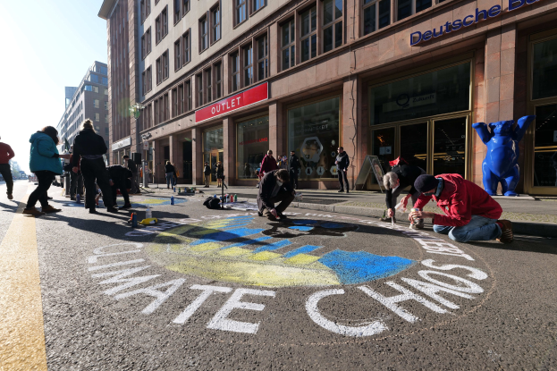Menschen sitzen vor einem Gebäude mit Fenstern und Namensschildern auf dem Boden, umgeben von Flaschen und anderen Gegenständen, mit Bäumen und einem klaren blauen Himmel im Hintergrund.