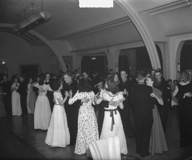 Schwarzes und weißes Bild von Menschen, die in einem Ballsaal tanzen, mit Deckenbeleuchtung umgeben von Stühlen.