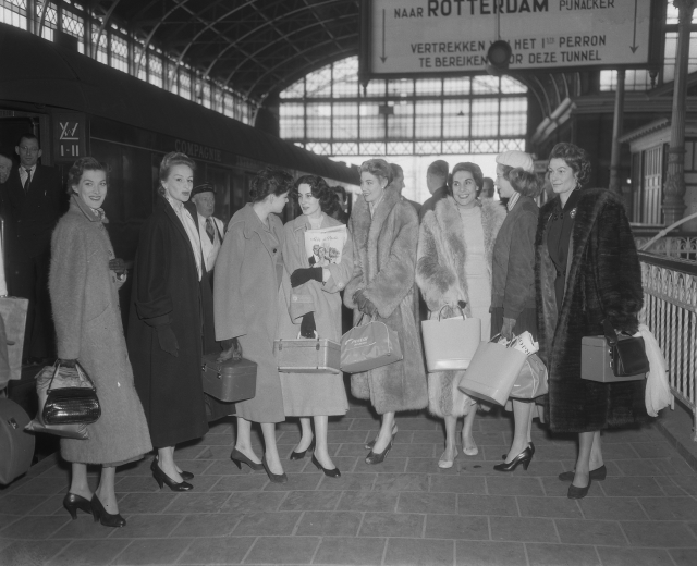 Schwarz-weißes Bild von Frauen an einem Bahnhof mit Taschen, mit einem Zug auf der linken Seite und Geländern und einer Tafel auf der rechten Seite.