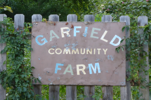 Ein Holzzaun mit einem Schild, auf dem 'Garfield Community Farm' steht, vor üppiger grüner Vegetation und Bäumen.