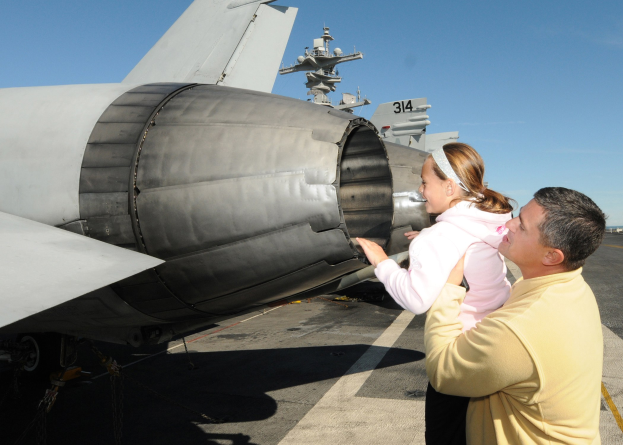 Ein Mann in einem gelben Hemd und schwarzen Hosen steht neben einer Frau, die ein Baby auf dem Flugdeck eines Flugzeugträgers hält, mit anderen Flugzeugen und einem klaren blauen Himmel im Hintergrund.