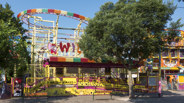 Freizeitpark mit Achterbahn in der Mitte, umgeben von Bäumen, Geländern, Pfählen, Schildern und anderen Strukturen, einer Straße unten und einem Himmel im Hintergrund.