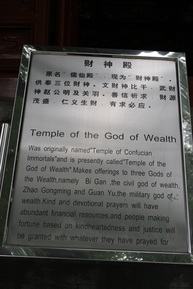 Ein Tempel, der dem Gott des Reichtums gewidmet ist, mit einer Namensplakette im Vordergrund und einer T├╝r im Hintergrund, umgeben von üppiger Vegetation.