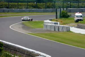 Ein Rennwagen fährt eine Rennstrecke entlang mit einem Polizeiwagen im Hintergrund, umgeben von Gras, Pflanzen, Bäumen und Zaun, mit einem Fahrzeug auf der rechten Seite und Text unten.