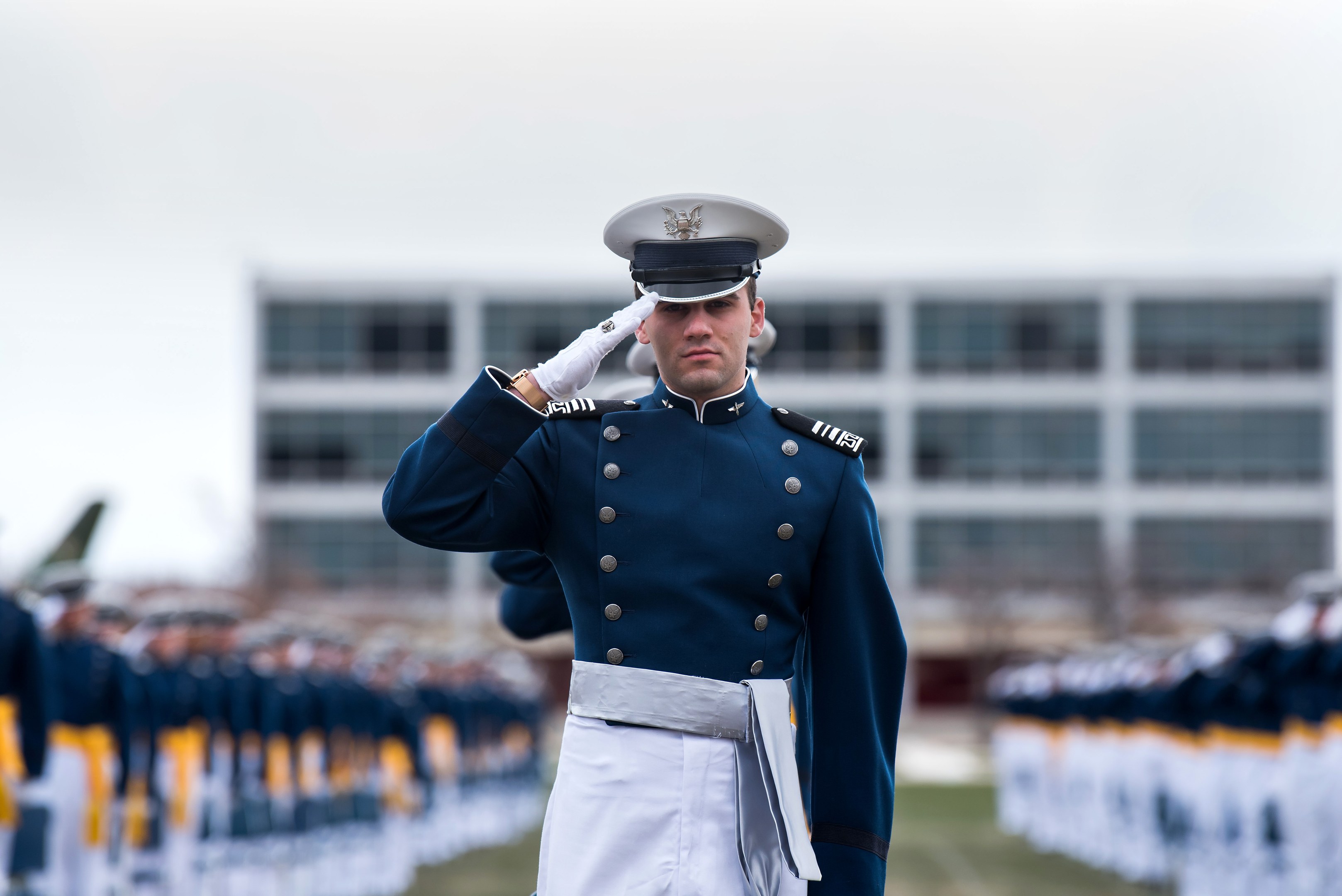 Ein Mann in militärischer Uniform salutiert auf einer Abschlussfeier, umgeben von einer Gruppe von Menschen auf dem Boden, mit einem Gebäude und Himmel im Hintergrund.