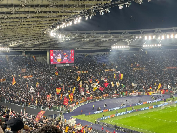 Eine große Menge im Stadion schwenkt Fahnen, mit Bannern auf dem Boden, Menschen in Mützen in den Rängen und einer beleuchteten Decke, was die Vorfreude auf ein Fußballspiel vermuten lässt.